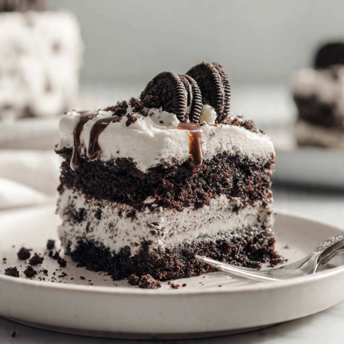 Oreo poke cake slice on cream plate showing dark chocolate cake layers, white Oreo cream topping, and chocolate drizzle