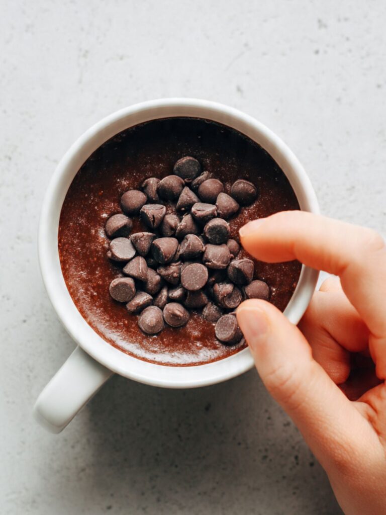 Fingertip pressing a cluster of semi-sweet chocolate chips just below the surface of chocolate mug cake batter in a white ceramic mug