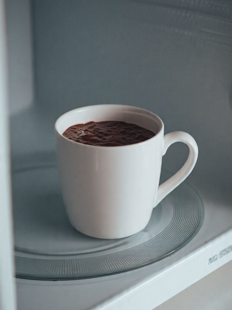 White ceramic mug with dark chocolate cake batter sitting centered on a white microwave glass turntable ready to cook
