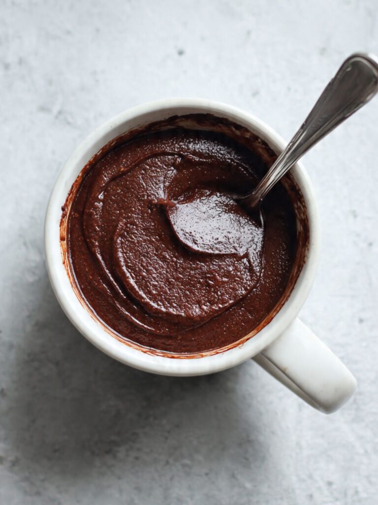 Thick dark glossy chocolate mug cake batter fully mixed in a white ceramic mug with a silver spoon resting across the rim