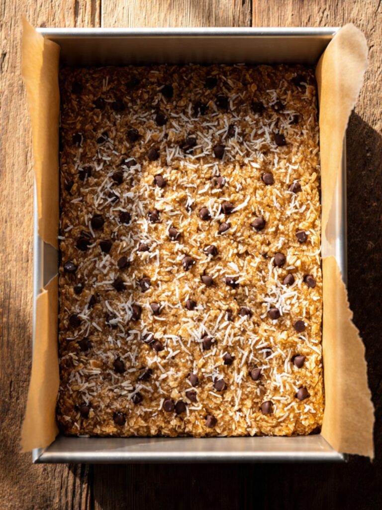 Coconut toffee bar batter pressed into parchment lined 9x13 pan with coconut and chips pressed on top