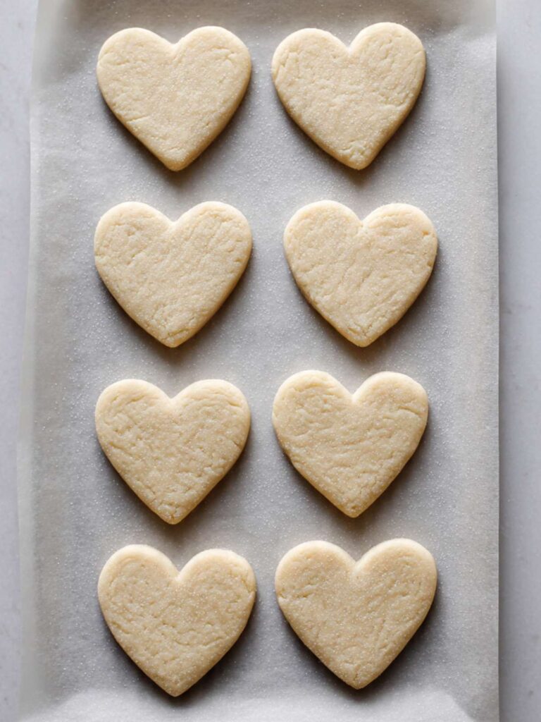 Unbaked heart-shaped and square sugar cookies arranged on a parchment-lined baking sheet before going into the oven, raw dough pale and smooth