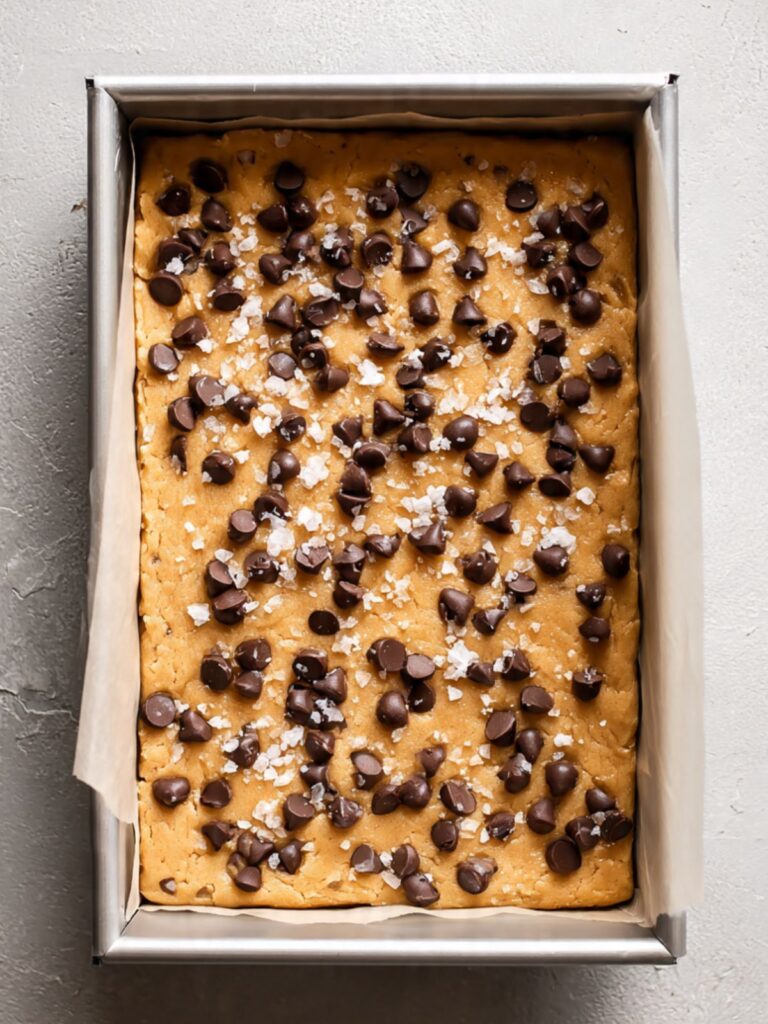 unbaked Nestlé Toll House cookie bar dough pressed into a parchment lined aluminum pan with chips and flaked salt on top