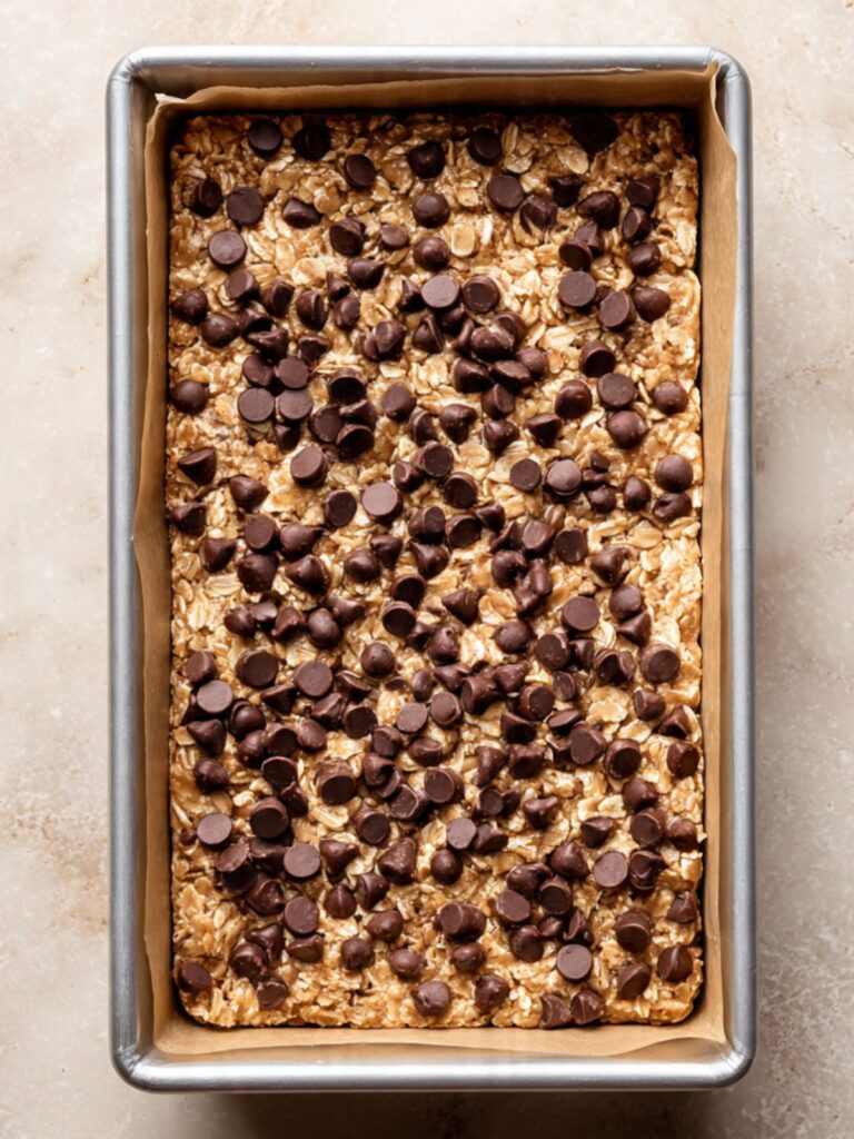 unbaked oatmeal chocolate chip bar batter pressed evenly into a parchment-lined 9x13 pan with semi-sweet chocolate chips scattered and pressed across the surface