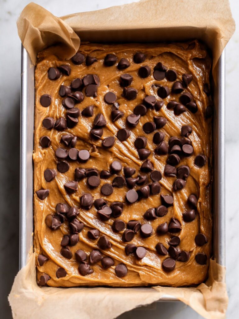 Overhead of peanut butter chocolate chip bar batter spread evenly in parchment-lined pan with chocolate chips scattered on top