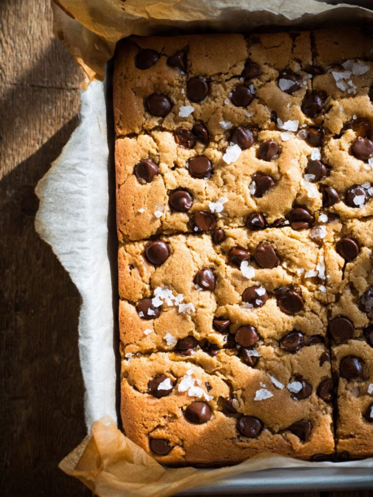 Freshly baked salted caramel chocolate chip cookie bars in a parchment-lined square pan, matte golden top
