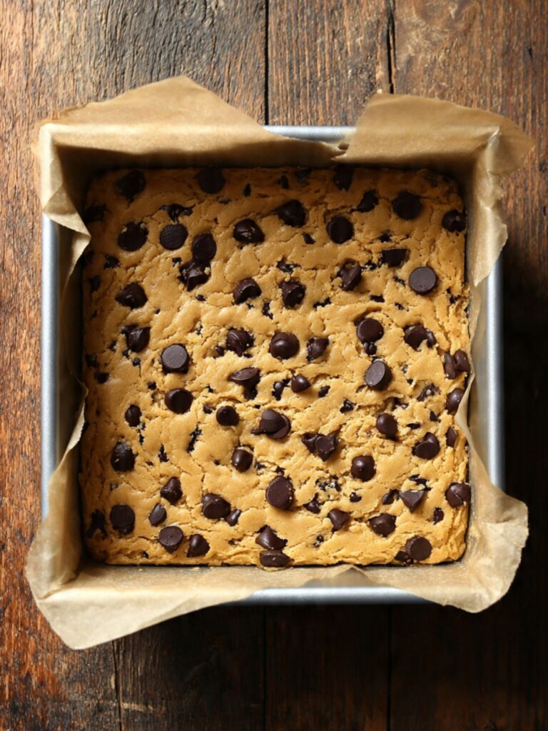 Thick chocolate chip cookie dough pressed into an even layer in a parchment-lined square silver baking pan, ready to bake